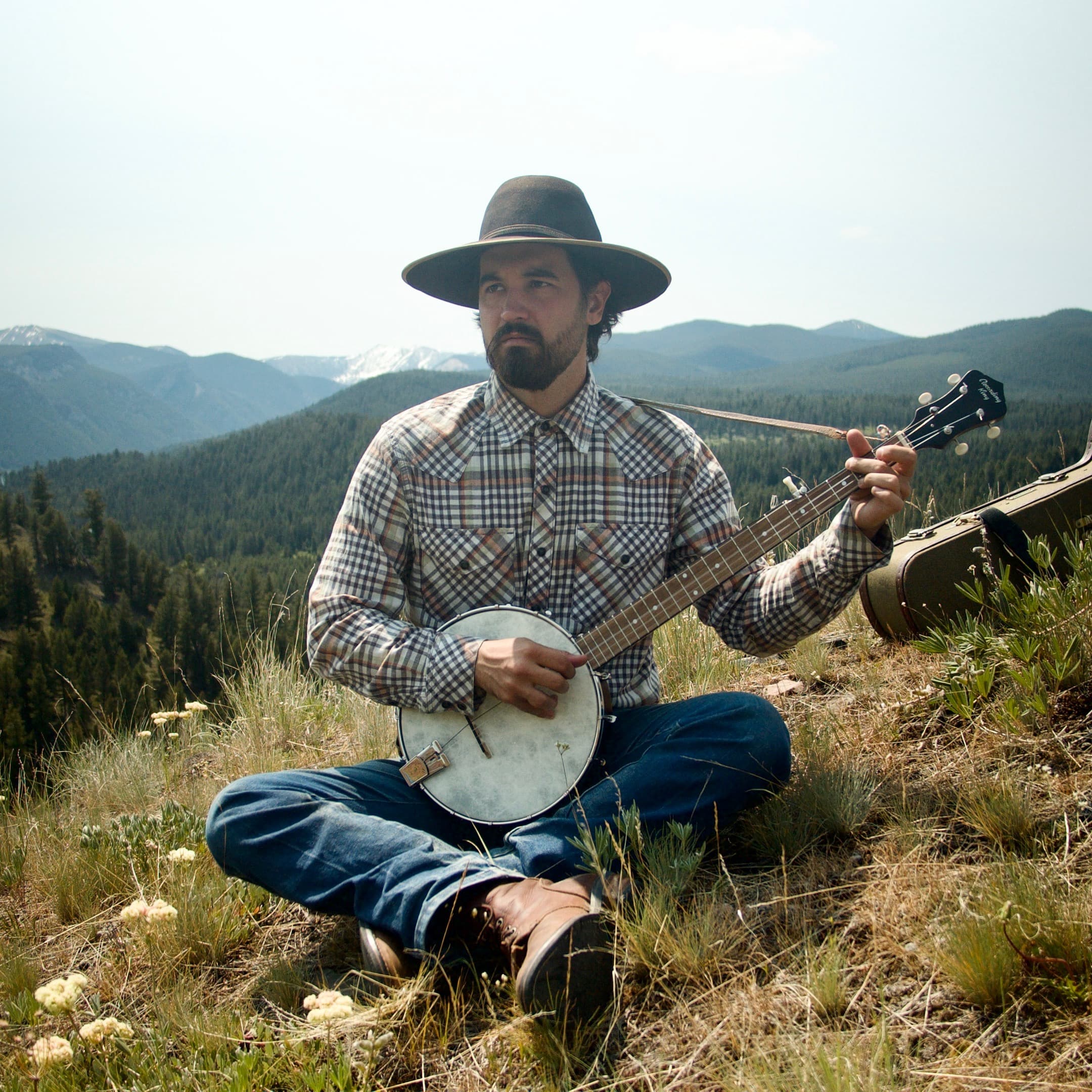 A man is wearing a hat and seating in a mountain setting and playing music