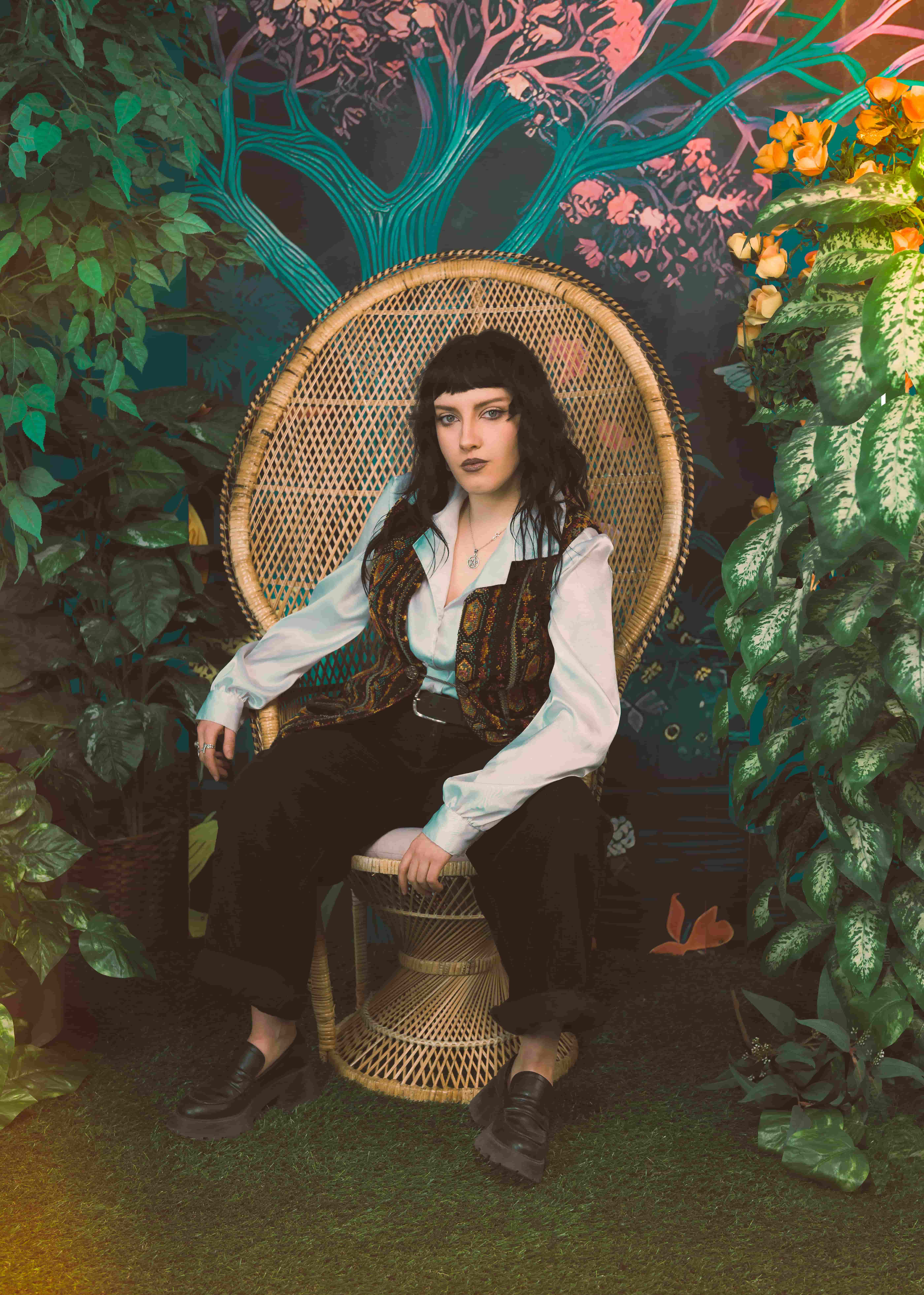A person seated in a woven wicker chair surrounded by lush green plants and a colorful mural backdrop. They wear a white button-up shirt, patterned vest, black pants, and black shoes, with dark hair and dramatic makeup, posing confidently in a stylized indoor garden setting
