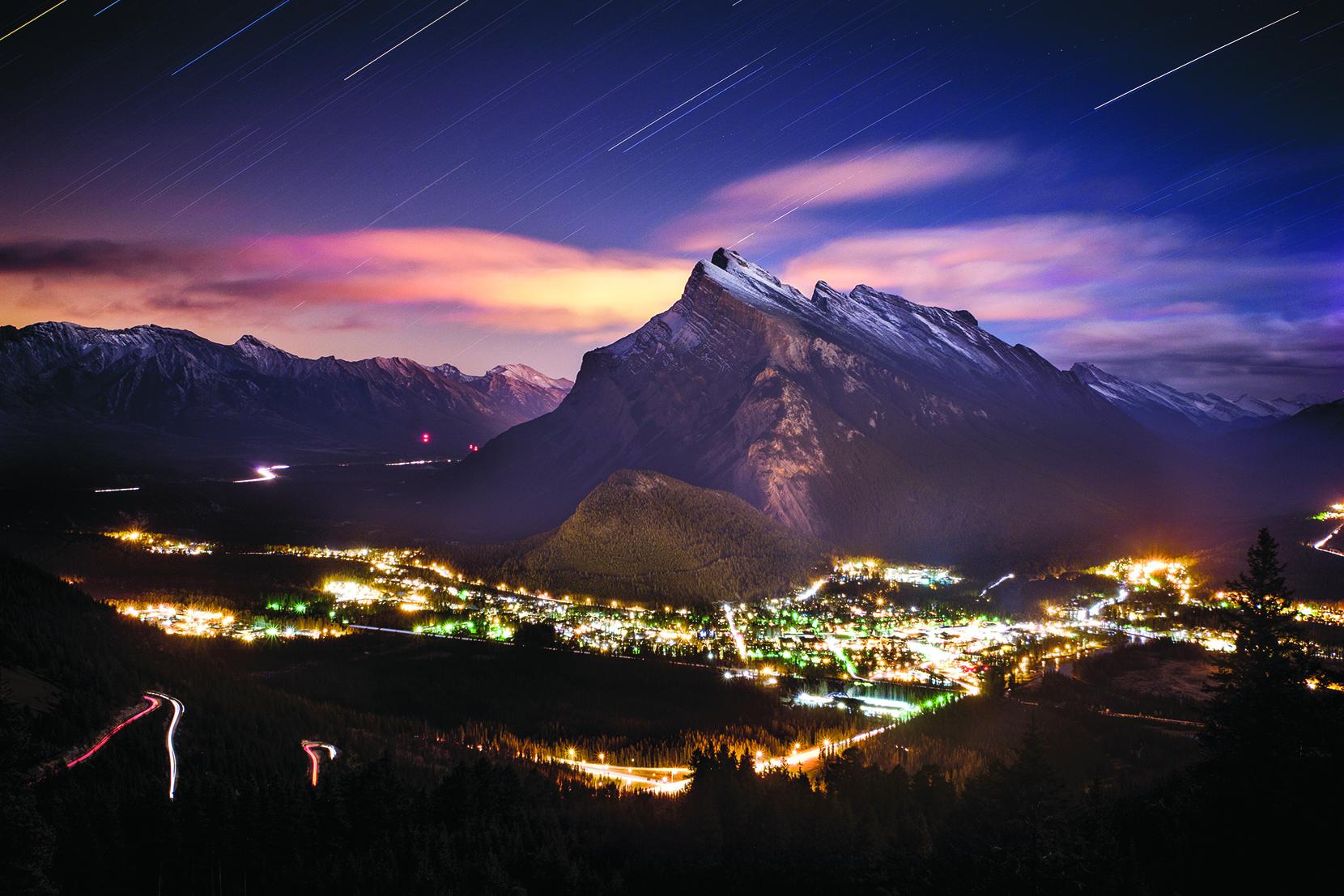 The Banff Townsite is illuminate at night. Stars wheel over Mt. Rundle and behind it effervescent clouds are lit up in this long exposure image.