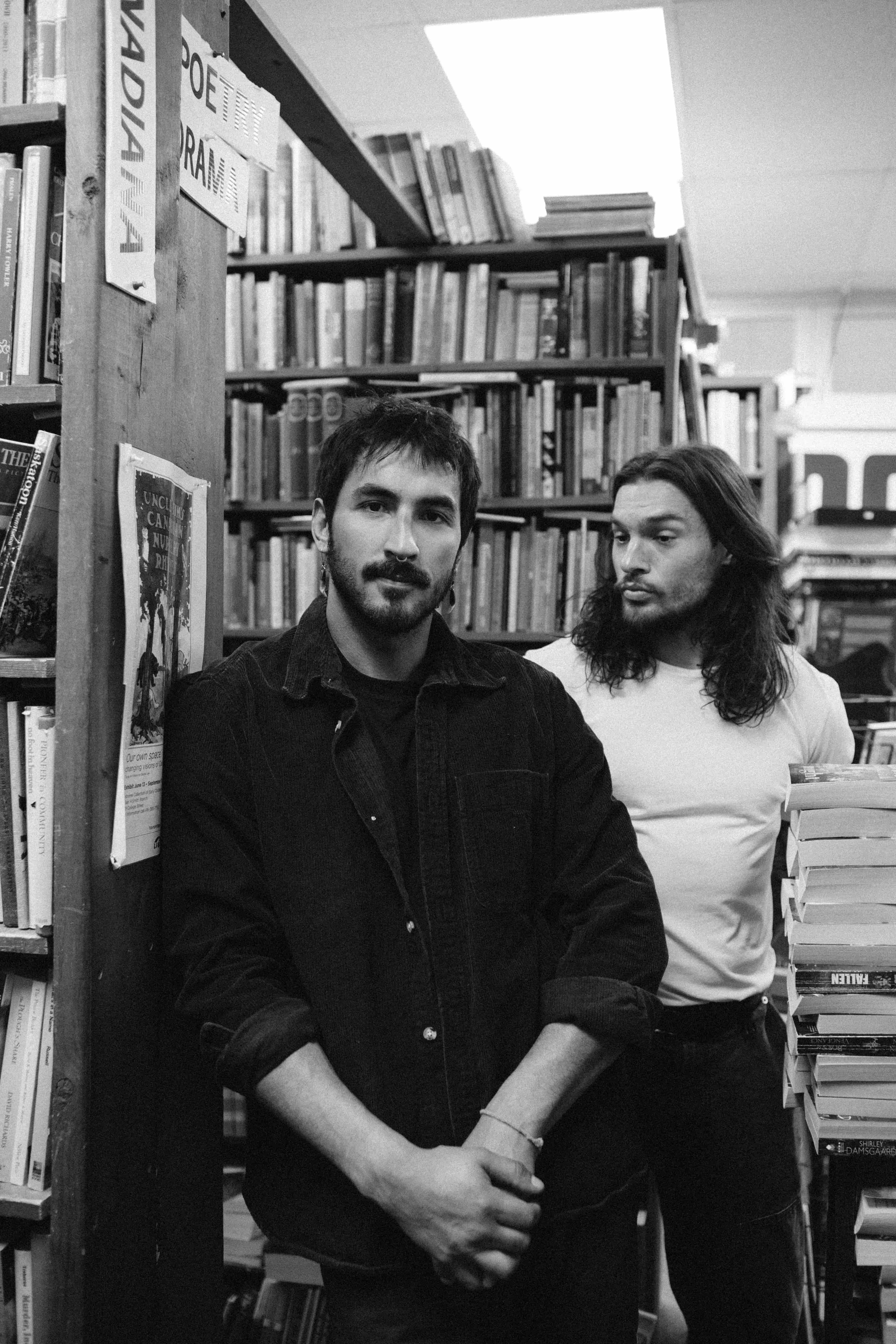 Two people standing between tall bookshelves in a bookstore or library, photographed in black and white. One person in the foreground wears a dark jacket and looks toward the camera, while the other stands behind beside a stack of books, creating an intimate, artistic portrait among rows of books.