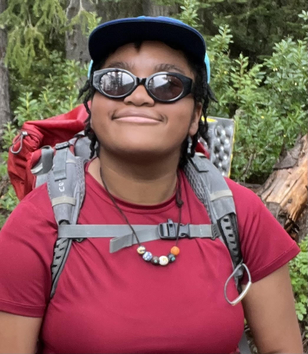 Léa Flowers Nelson in sunglasses and backpack smiles, with trees behind