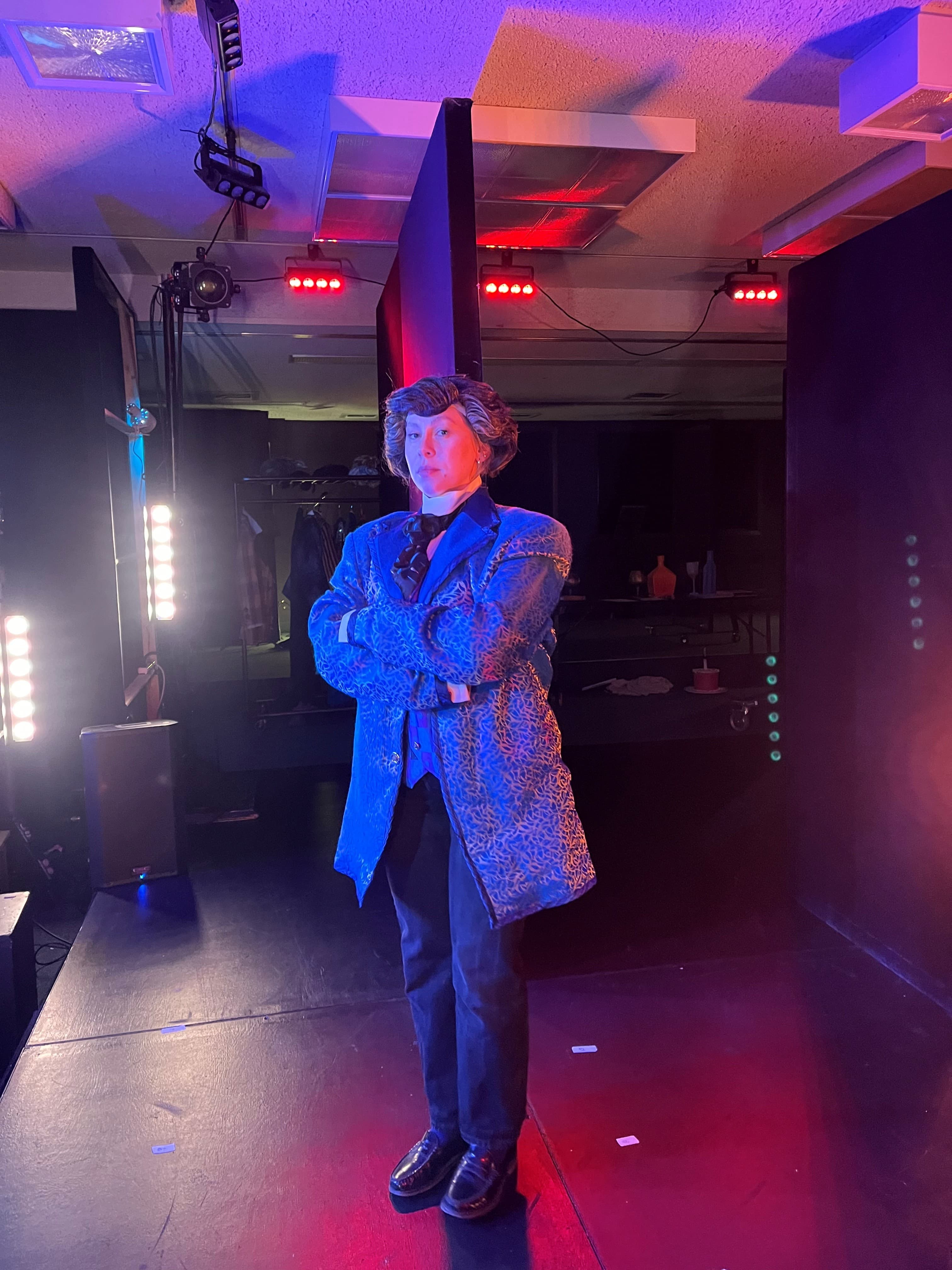 A person stands confidently on a small stage with their arms crossed, wearing a patterned blue coat and dark outfit under dramatic red and blue lighting. The theatrical setting, stage lights, and bold pose create a striking backstage or performance atmosphere.”