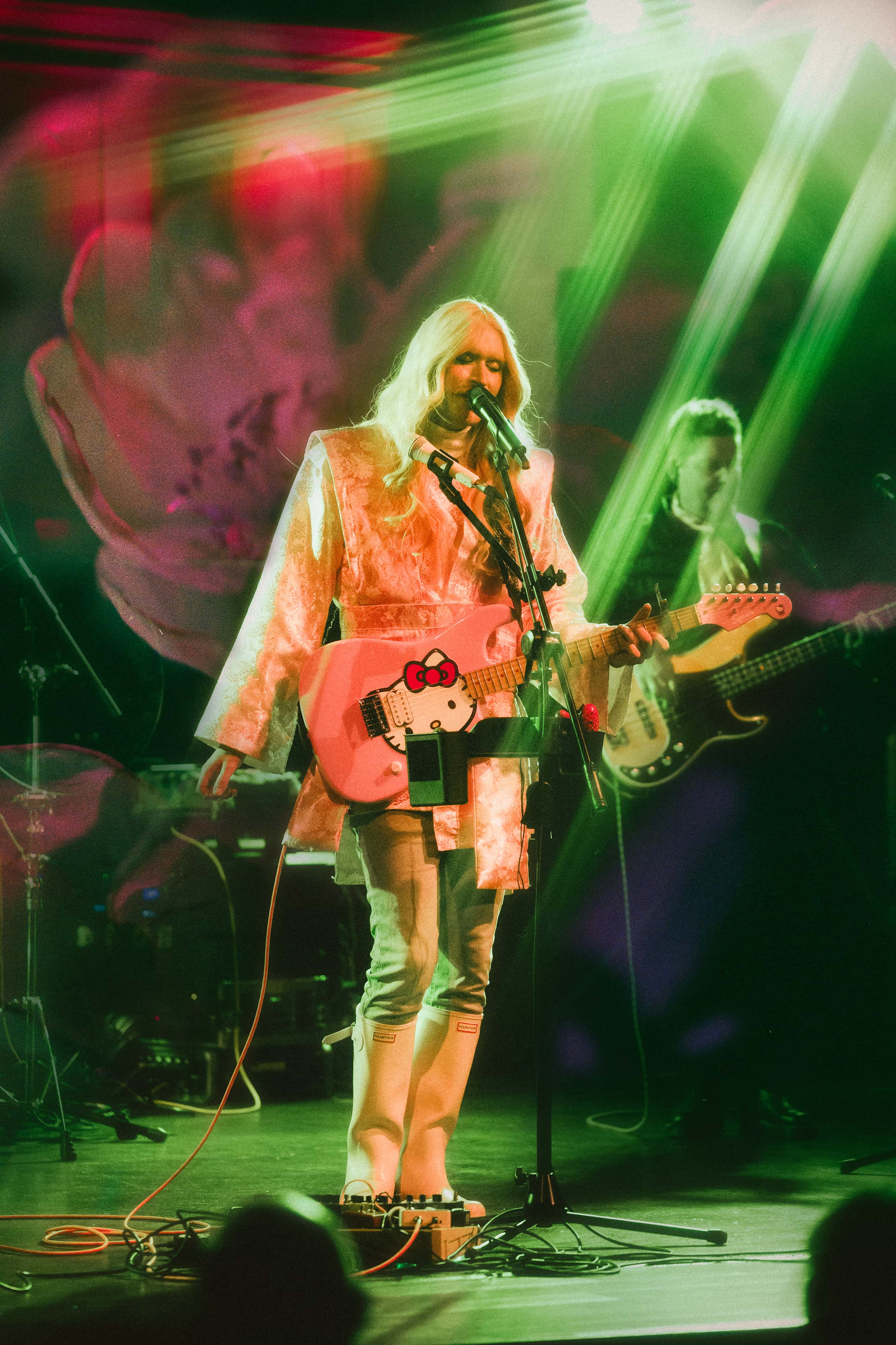 A musician with long blonde hair performs on stage under colorful green and pink lighting, singing into a microphone while playing a pink Hello Kitty electric guitar. Another band member is visible in the background, creating a dreamy, energetic live concert atmosphere.