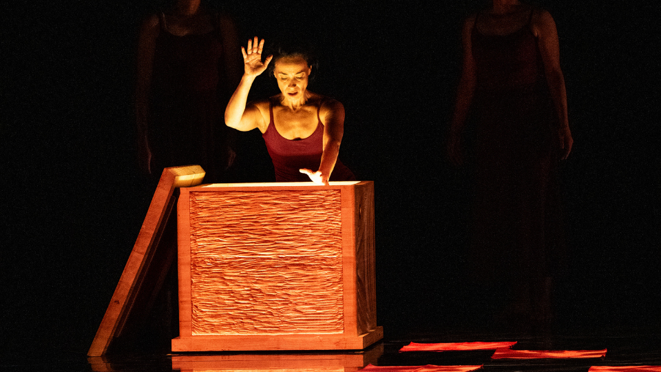 Woman on stage is facing a large wooden box with her right hand lifting on the air