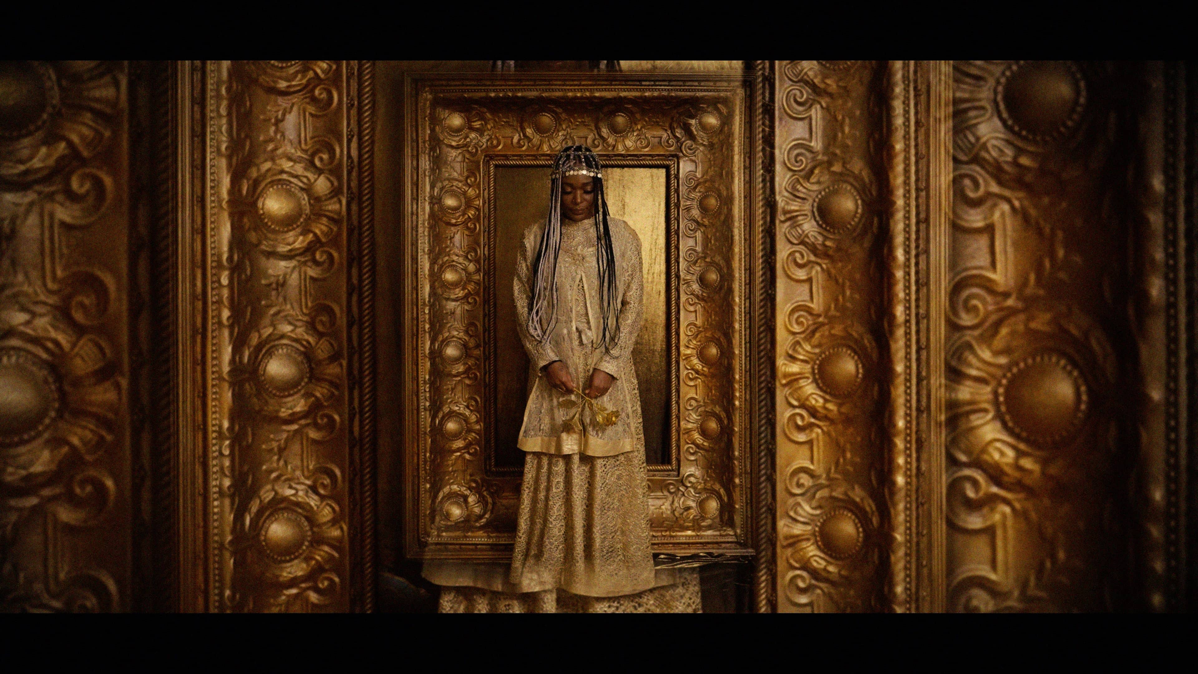 A solitary figure stands centered within an elaborate gold architectural frame, creating a dramatic and almost sacred composition.