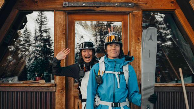 Two women with ski gears are standing in a front of a contemporary chalet door. The snowy high peaks, mountains and trees are reflecting into the windows 