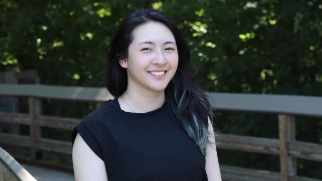 Smiling woman with dark hair posing by a wooden fence.