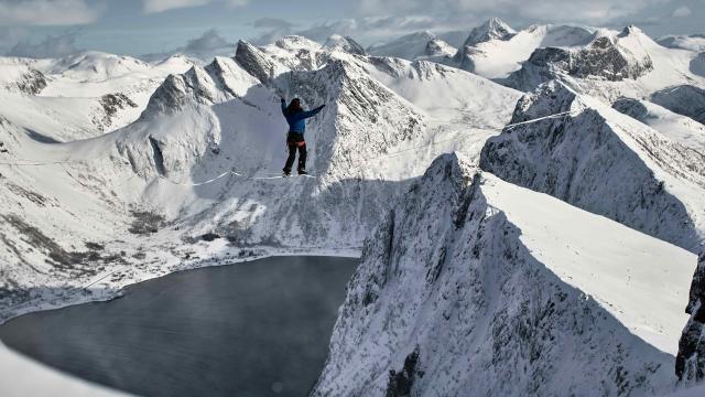 An athlete is walking on a slackline amongst snowcapped mountain peaks.