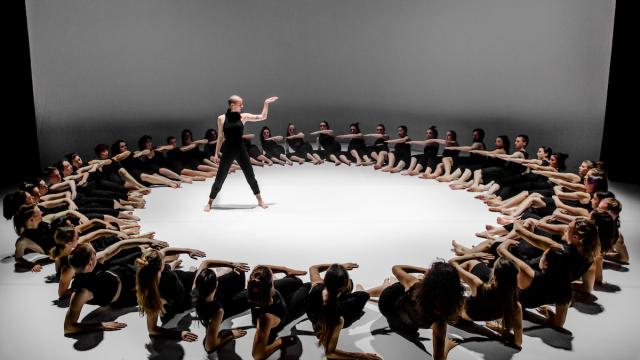 50 dancers dressed in black form a circle around a single dancer standing in the center