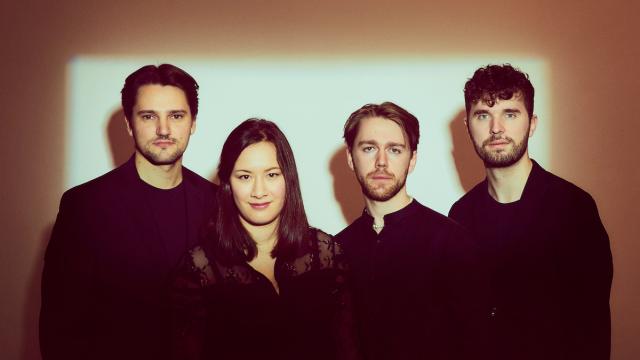 Four musicians dressed black, standing in front of a backlit wall