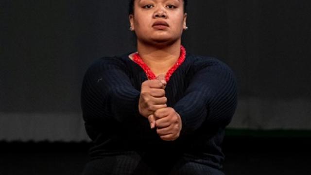 An indigenous woman sits on the ground with her hands in stacked fists held in front of her. 