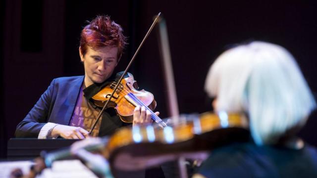 Two women face each other playing violin, the one head-on from the photographer is in focus.