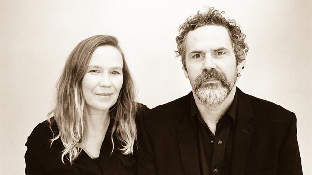 Pharis and Jason Romero stand shoulder to shoulder smiling in black dress clothes.