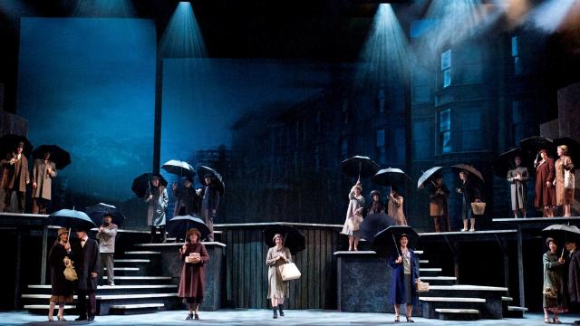 A stage set to look like an early 20th century street is full of individuals on a multi-tiered stage holding umbrellas. 