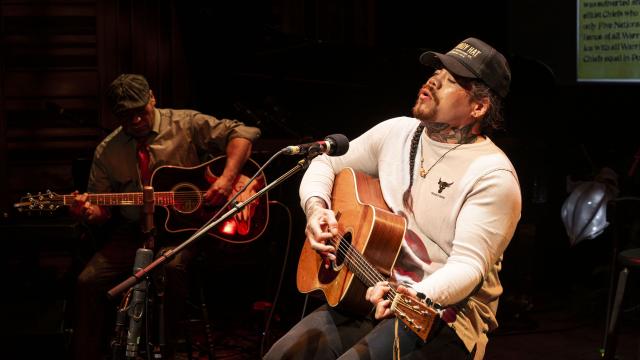 A musician plays the guitar while singing into a microphone with another guitarist in the background