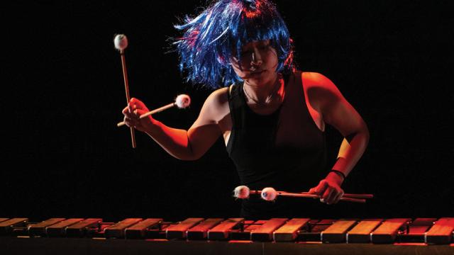 A musician in a blue wig plays a percussion instrument
