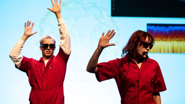 Two musicians in bright red coveralls perform in front of a blue backdrop