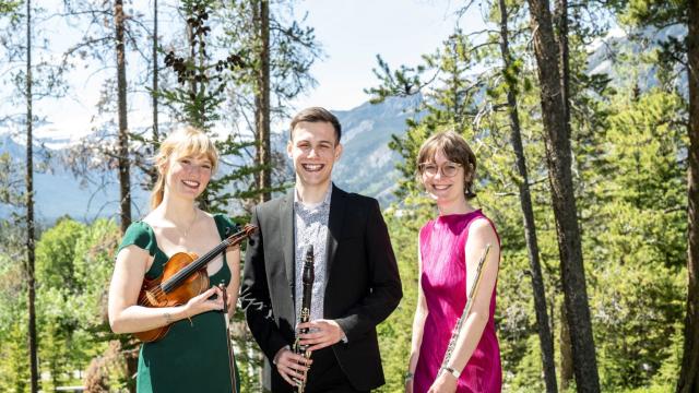 Woman in green dress holding a violin standing beside man in suit with a clarinet in front of him next to a woman in a pink dress holding a flute casually at her side