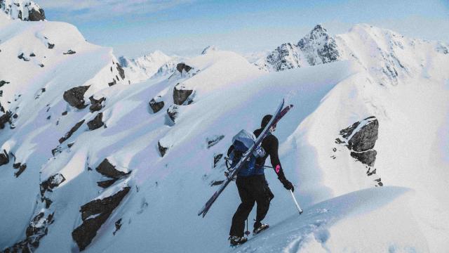 Alpine skier ascends a steep, snowy mountain ridge with tools and skis strapped to their backpack. 