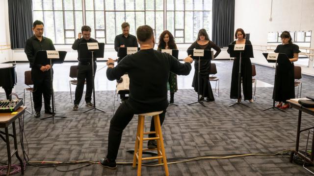 A row of opera singers stand next to music stands during a rehearsal in a large studio