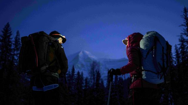 In a dark setting, a man and women with warm winter clothes, backpack, headlamp and climbing gear looking at each other, facing a snowy mountain 