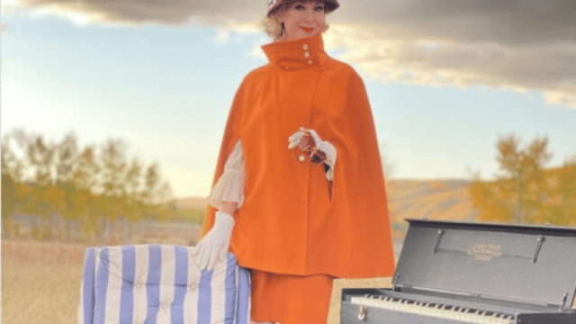 Performer in a striking vintage-inspired orange outfit poses beside an outdoor piano, creating a theatrical scene against a dramatic prairie landscape.