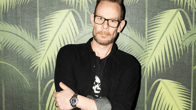 Portrait of a DJ standing confidently with arms crossed in front of a tropical palm leaf backdrop.