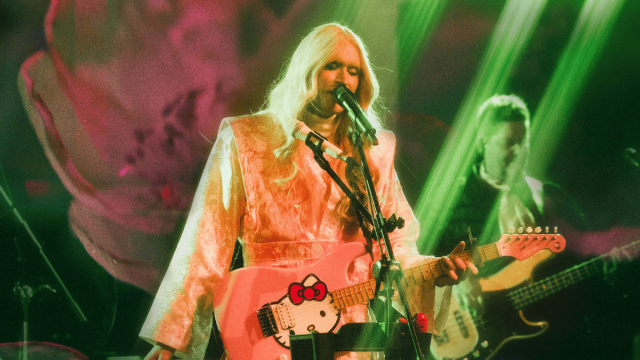 Performer with long platinum-blonde hair sings into a microphone while playing a pink electric guitar under green stage lights, with another musician performing in the background.
