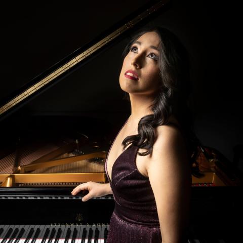 Woman with long dark hair standing in front of grand piano in formal wear looking dramatically up and off camera