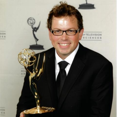 Profile of Dave Pierce in a black suit holding a Emmy award 