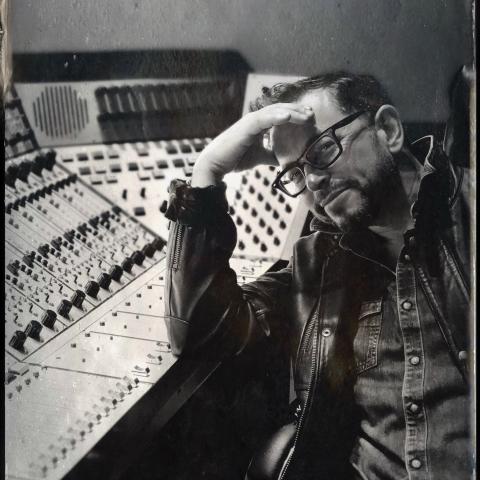black and white portrait of a man with glasses seating next to a mixing console.