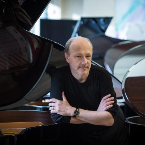 Man in black shirt sitting with arms crossed in front of several grand pianos