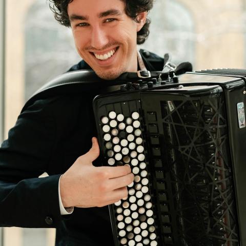 Michael Bridge is playing a black accordion and smiling at the camera