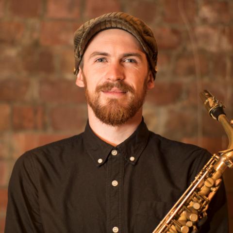Man wearing cap and black button up smiling at camera and holding a saxophone in front of him