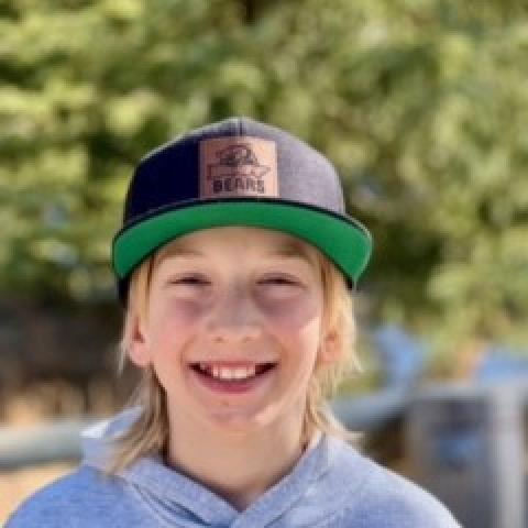 Young blonde boy smiling directly at camera outside wearing a baseball cap