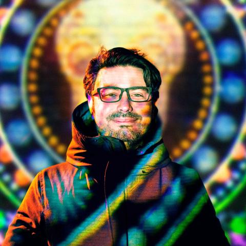 A man wearing glasses and a winter coat standing in front of a very colourful projection on a wall which covers his body
