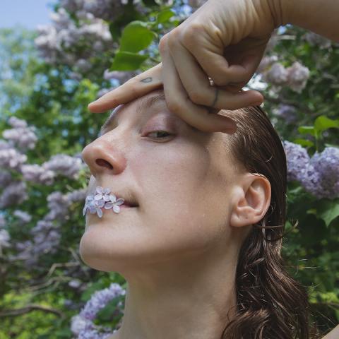 Lucy has little purple flowers in their mouth. Their hand is resting on their face above their eyebrow. They have long h