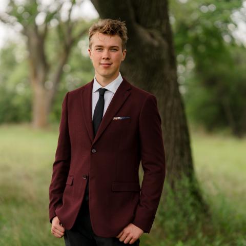Wide shot of man wearing a tie and a burgundy suit standing in a field with trees