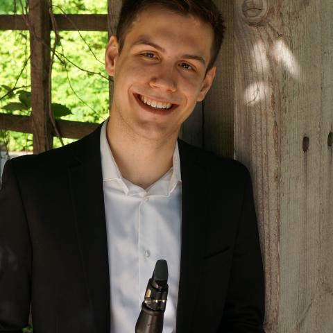 Man smiling directly at the camera holding his clarinet and wearing a dark suit jacket with white shirt