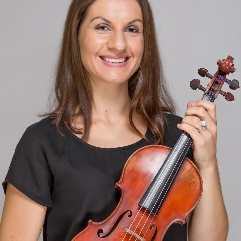 Woman holding a violin facing outward to the camera with a large smile looking directly at the camera