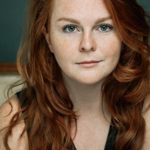 Close up photo of a red haired woman with long flowing wavy hair smiling slightly at the camera