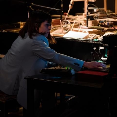 Anoush sits at a piano with additional musical electronic equipment. Anoush wears a grey blazer. There is light on the p