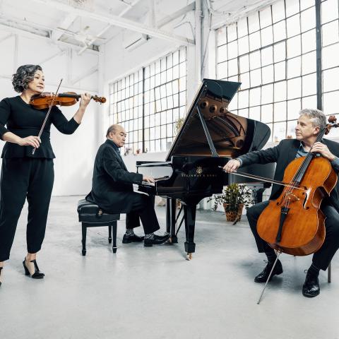 Annalee, Jamie, and Roman (left to right) are sitting in a modern white room playing their instruments (violin, piano, a