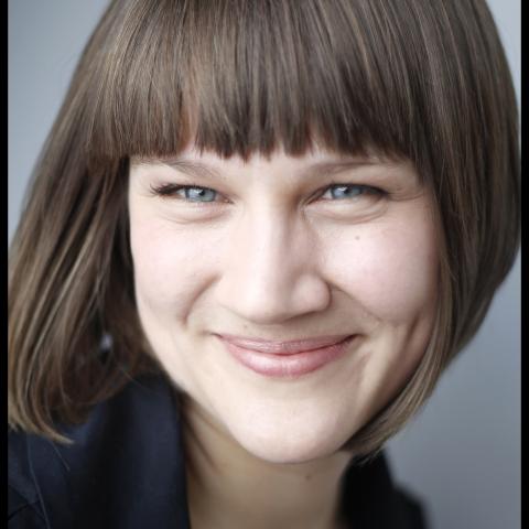 A close up shot of Madeline. She is smiling and wears a blue shirt. The background is grey. 