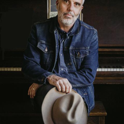 John sits on a piano bench with the piano behind him. His arms are crossed and he holds a grey hat. He looks at the came