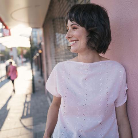 Erin stands outdoors on a sidewalk in front of a dusty rose wall. Erin looks to the left and is smiling. 