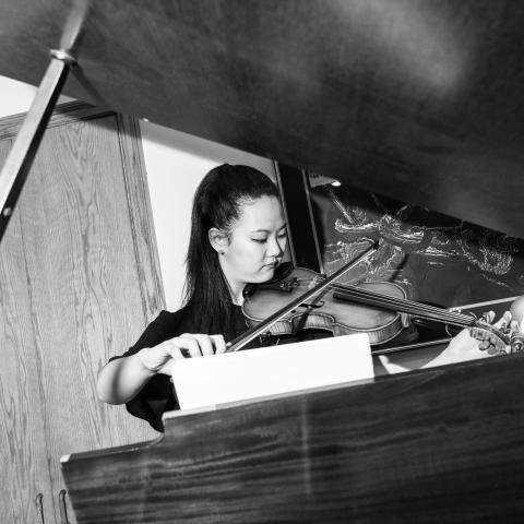A black and white photo of Katherine playing a violin. She stands in front of a piano and the photo is shot through the 