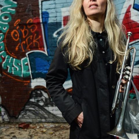 Lina stands with a trumpet in her hand. She stands against a colourful brick wall.