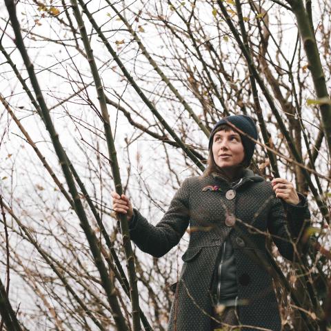 Myriam is outdoors in shrubby trees wearing a winter coat. It looks cold and damp but she looks happy. 