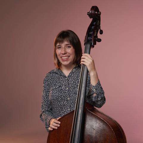 Zoe stands with a patterned shirt holding an upright bass in front of a red background. 